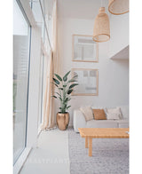 Un salon lumineux et moderne avec de grandes fenêtres, un canapé blanc, des coussins neutres, une table basse tressée, deux œuvres murales abstraites, une plante Heliconia artificielle de 180 cm dans un pot structuré et des suspensions tressées suspendues au plafond.