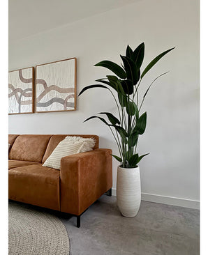 Un salon moderne avec un canapé en cuir marron, des coussins rayés et la plante artificielle Heliconia 180 cm dans un vase blanc. Au-dessus, des œuvres d'art abstraites sont accrochées sur un mur blanc avec un sol gris et un tapis beige - idéal pour s'inspirer de l'achat de plantes artificielles.
