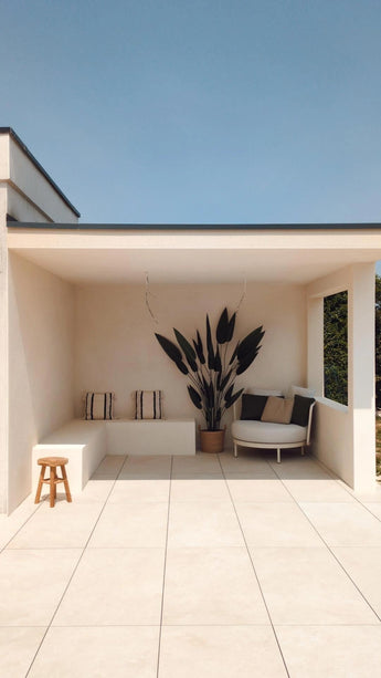 Une terrasse moderne et minimaliste avec du carrelage couleur crème, un canapé intégré avec coussins rayés, une plante artificielle Heliconia 210 cm pour une touche luxueuse, un petit tabouret en bois et une chaise ronde avec coussins sous un toit plat sur un ciel bleu clair.