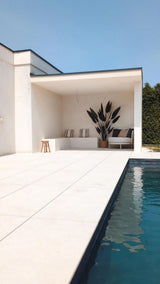 Piscine extérieure moderne avec carrelage clair au sol, un coin salon recouvert de blanc avec des coussins rayés, une plante artificielle Heliconia de 210 cm pour une touche luxueuse, un petit tabouret, un canapé, un ciel bleu au dessus et une haie verte pour compléter le tableau.