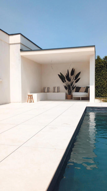 Piscine extérieure moderne avec carrelage clair au sol, un coin salon recouvert de blanc avec des coussins rayés, une plante artificielle Heliconia de 210 cm pour une touche luxueuse, un petit tabouret, un canapé, un ciel bleu au dessus et une haie verte pour compléter le tableau.