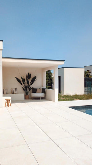 Une terrasse extérieure moderne avec du carrelage blanc au sol, des sièges ombragés au look luxueux, la plante artificielle Heliconia de 210 cm comme pièce maîtresse, des coussins douillets, un tabouret en bois et une piscine au premier plan sous un ciel bleu clair.