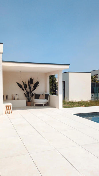 Une terrasse extérieure moderne avec du carrelage blanc au sol, des sièges ombragés au look luxueux, la plante artificielle Heliconia de 210 cm comme pièce maîtresse, des coussins douillets, un tabouret en bois et une piscine au premier plan sous un ciel bleu clair.