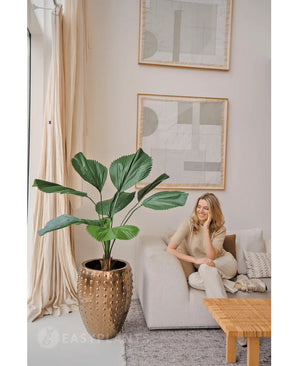 Une femme se détend sur un canapé lumineux dans un salon moderne aux couleurs neutres. À côté d'elle se trouve la plante artificielle Lucuala Grandis de 120 cm dans un pot, tandis que des œuvres d'art abstraites sont suspendues au-dessus et que la lumière naturelle illumine l'espace.