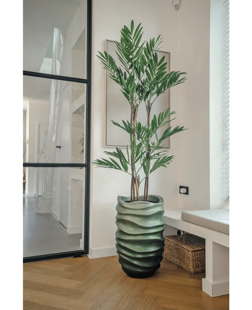 La plante artificielle Parlour Deluxe 120 cm a des tiges fines et de longues feuilles vertes dans un vase vert ondulé, parfait pour les pièces modernes et lumineuses avec des parquets en bois clair et de grandes fenêtres.