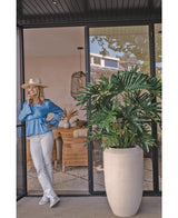 Une femme vêtue d'un chemisier bleu, d'un pantalon blanc et d'un chapeau rit devant une porte vitrée à côté de la plante artificielle Philodendron de 125 cm dans un pot beige. Des chaises et une décoration en osier sont visibles dans toute la pièce.