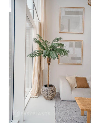 La Plante Artificielle Phoenix Palm De Luxe 180 cm en pot structuré est placée à côté de grandes fenêtres dans un salon lumineux au mobilier neutre, avec un canapé blanc, des coussins beiges, une table en bois et de l'art abstrait au mur.