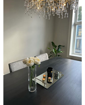 Une table à manger moderne avec des chaises grises, un bouquet de roses artificielles "Carmen" Real Touch Crème avec un bouquet de 36 cm, des bougies et un décor sur un plateau miroir. Un lustre est suspendu au-dessus, avec une fenêtre et une plante en pot en arrière-plan.