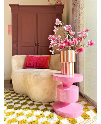 Un salon cosy avec un canapé rond beige, un coussin géométrique rose et orange, une table d'appoint rose, une branche artificielle Magnolia à petites fleurs 89 cm rose dans un vase, un tapis à motifs jaune-blanc et une armoire marron en fond.
