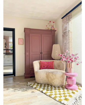 Un coin lecture confortable comprend une chaise crème, des coussins roses, un tapis à motifs jaunes et une armoire mauve. La Branche Artificielle Magnolia à petites fleurs 89 cm rose se dresse sur une table d'appoint rose, tandis que de grandes fenêtres remplissent cet espace invitant d'une lumière douce.