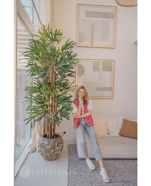 Une femme vêtue d'un cardigan à motifs roses et d'un jean sourit sur un canapé beige à côté du Lady Artificial Palm 210 cm dans un salon lumineux et moderne avec des œuvres d'art abstraites au mur.