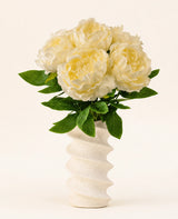 Un vase blanc ondulé et structuré contient le bouquet de fleurs artificielles en soie Peony White - de grandes fleurs de pivoines artificielles de couleur crème avec des feuilles vertes sur un fond clair uni.