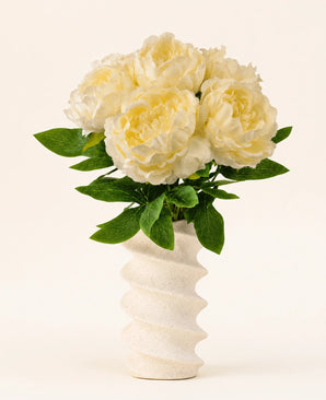 Un vase blanc ondulé et structuré contient le bouquet de fleurs artificielles en soie Peony White - de grandes fleurs de pivoines artificielles de couleur crème avec des feuilles vertes sur un fond clair uni.