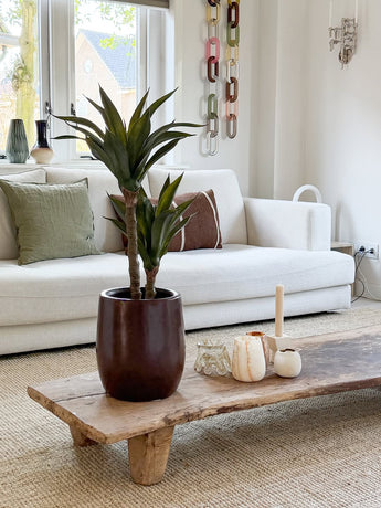 Un salon moderne avec un canapé blanc, des coussins verts et marron, une table basse en bois décorée de bougies et d'agave artificielle 76 cm, et une décoration murale en maille chaîne. La lumière naturelle pénètre par les fenêtres derrière le canapé.
