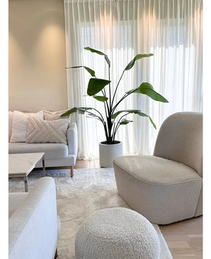 Salon lumineux et moderne avec un canapé blanc, un fauteuil rond et un pouf. Une plante artificielle Strelitzia Real Touch de 180 cm prête à l'emploi dans un pot beige se trouve à côté de rideaux transparents. Un décor neutre et confortable avec un tapis moelleux et des coussins complètent le look.