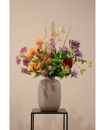 Un vase beige avec branche de renoncule artificielle x5 Real Touch orange 62 cm de renoncule orange artificielle se dresse sur un support en métal noir sur un fond clair uni.