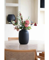 Le Vase Artic Noir Ø21 x H29 cm, rempli de fleurs roses, blanches et rouges, repose sur une table ronde beige entourée de deux chaises en bois aux dossiers en osier tressé. Des planches décoratives sont visibles en arrière-plan.