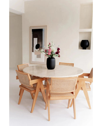 Une table ronde en marbre avec quatre chaises en bois entoure le Vase Artic Noir Ø21 x H29 cm rempli de fleurs. Des œuvres d'art murales abstraites et une pièce de décoration noire sur une étagère intégrée complètent la pièce minimaliste aux tons clairs.