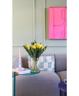 Le bouquet artificiel en soie Tulipes dans un vase Jaune se trouve sur une table d'appoint verte à côté d'une bougie torsadée rose, de deux livres et d'un canapé gris structuré avec un coussin à motifs. Une œuvre d’art abstraite rose vif est accrochée au mur vert clair.