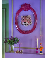 Un mur violet vif présente une tête de léopard décorative dans un cadre orné de rose. Ci-dessous, vous voyez le bouquet artificiel de tulipes en soie dans un vase violet, à côté d'une bougie violette, d'un chandelier doré et de bougies colorées sur une étagère.