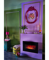 Un salon moderne avec des murs verts, une cheminée violette et des œuvres d'art léopard encadrées de roses. Un panier avec une couverture, un bouquet artificiel en soie, des tulipes dans un vase violet et des bougies décorent la pièce. Des livres et une lampe rose sont posés sur une table d'appoint vert-bleu.