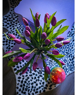 Le bouquet artificiel en soie Tulipes dans un vase Violet repose sur une table lavande à côté d'une boule disco vibrante sur un tapis à imprimé léopard noir et blanc.