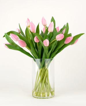 Le bouquet artificiel en soie Tulipes dans un vase Rose présente des tulipes roses avec de longues tiges et des feuilles vertes dans un vase en verre transparent, sur un fond blanc uni.
