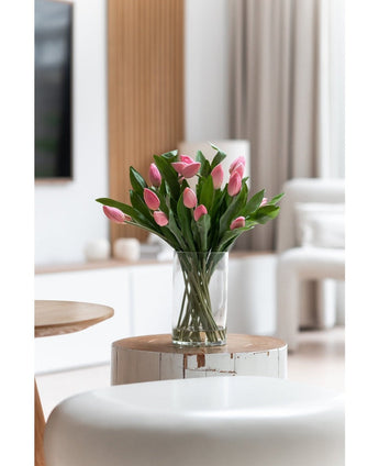 Le bouquet artificiel en soie Tulipes en vase Rose se dresse sur une table ronde en bois blanc dans un salon clair et moderne aux tons neutres et à la décoration contemporaine.