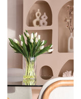 Le bouquet artificiel en soie Tulipes dans un vase Blanc, avec des tulipes blanches et des feuilles vertes, se dresse sur une table en verre. Derrière lui se trouvent des pièces de décoration modernes en céramique et une branche fleurie dans un vase blanc sur des étagères beiges.