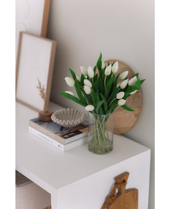 Le bouquet d'art en soie Tulipes dans un vase Blanc se trouve sur une table blanche à côté de livres empilés, d'une pièce de décoration en forme de coquille et d'œuvres d'art encadrées contre un mur de couleur claire.